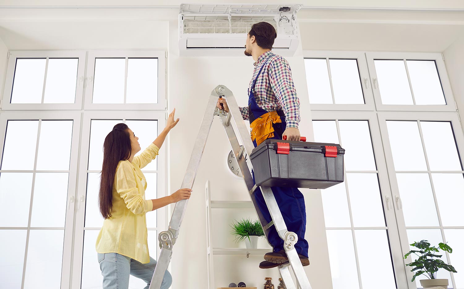 Woman showing an HVAC repairman standing on a ladder where their ductless unit is